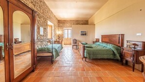 A room with a stone wall, a bed with a green bedspread, a wooden wardrobe, and a chair.