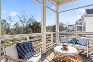 A covered outdoor deck with wicker furniture and cushions, a round table with a bowl, and a view of a house and trees.