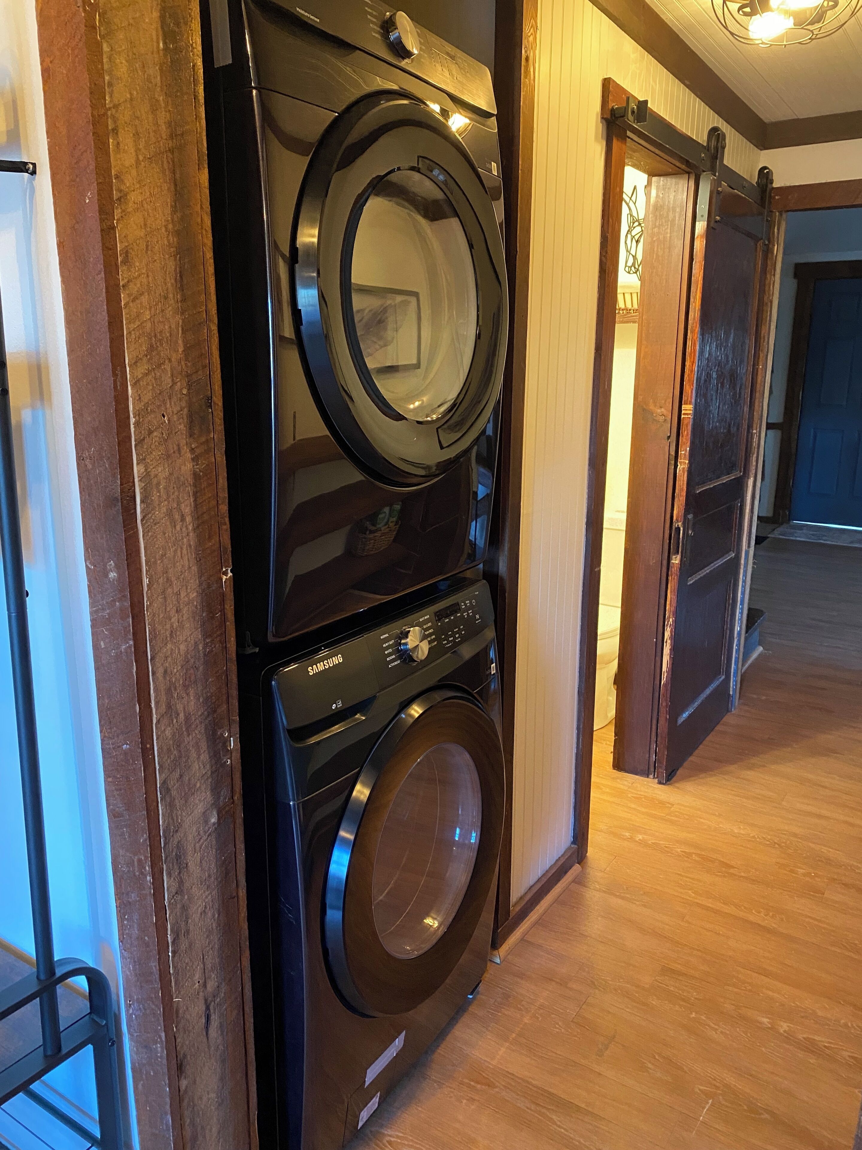 A stacked washer and dryer unit in a hallway with wooden flooring and a door leading to another room.