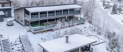 Un edificio moderno de varios pisos con techo plano y un área de estar al aire libre cubierta, sobre un paisaje cubierto de nieve.
