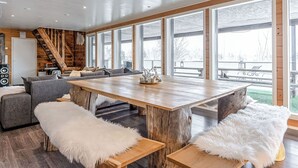 Ein geräumiger Raum mit einem großen Holztisch, grauen Sofas, weißen, flauschigen Hocker und großen Fenstern mit Blick ins Freie.