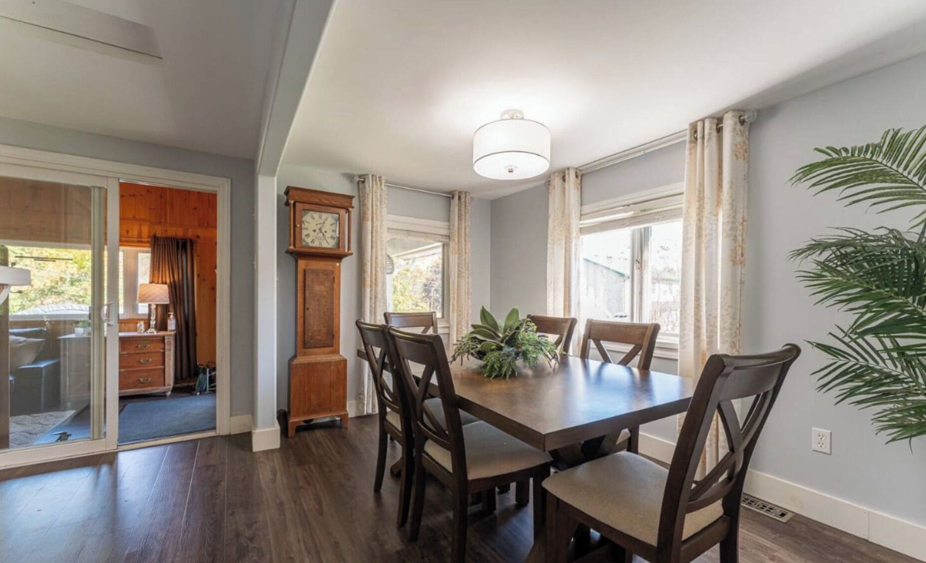 A dining area with a wooden table and chairs, a large clock, and a view of the outdoors.