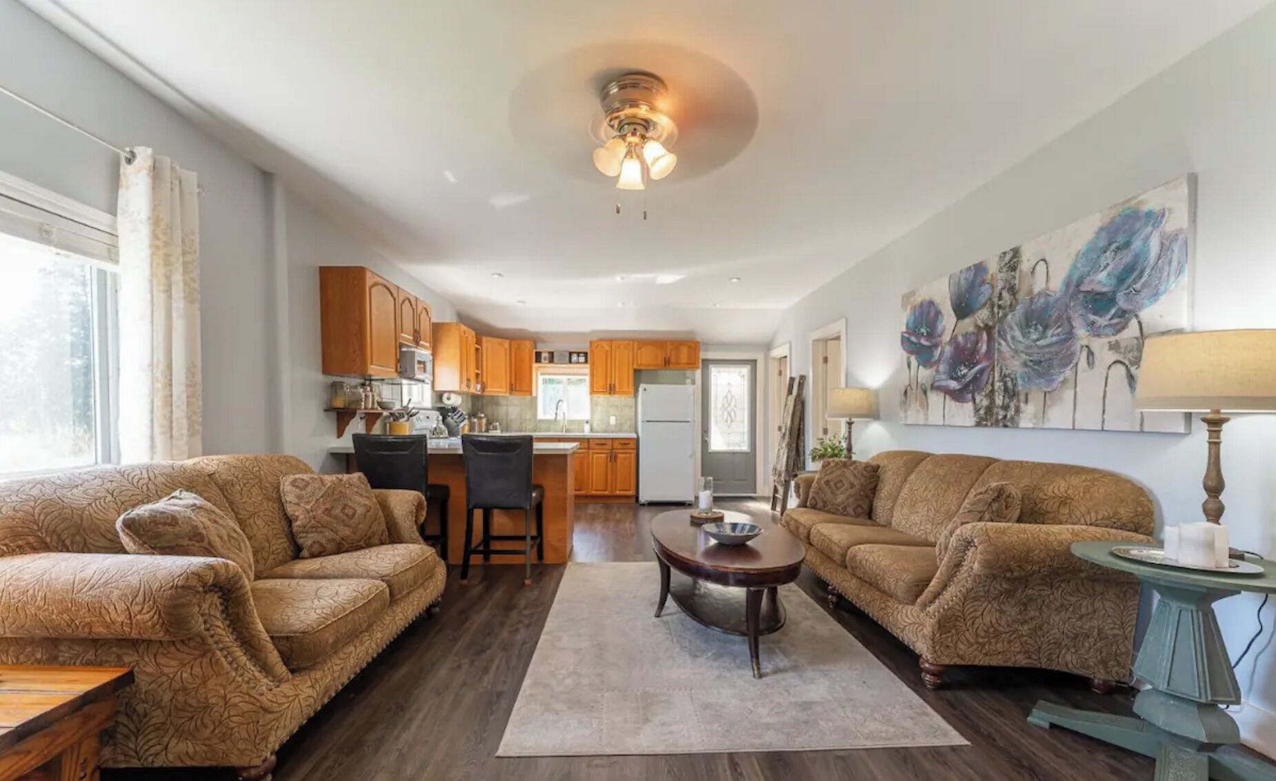 A spacious living room with two sofas, a coffee table, and a dining area with chairs. The kitchen is visible in the background with wooden cabinets and a refrigerator.
