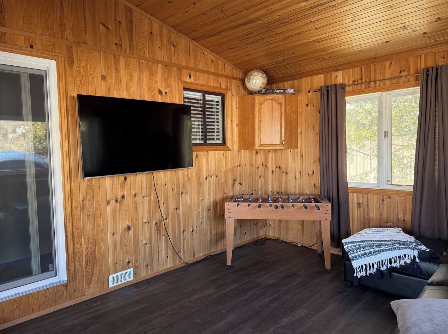 A cozy room with wooden walls, a flat-screen TV mounted on the wall, a foosball table, a sofa, and a window with curtains.