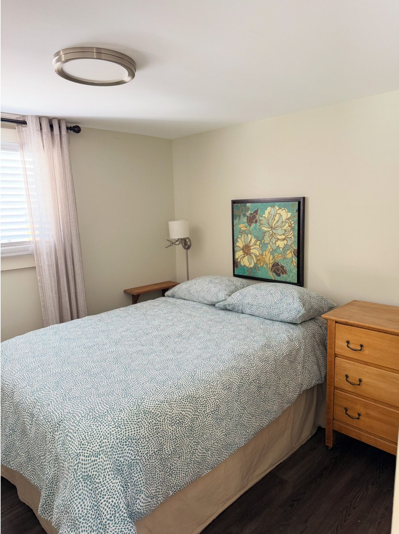 Un dormitorio con cama, mesita de noche, lámpara, cómoda de madera y un cuadro floral en la pared.