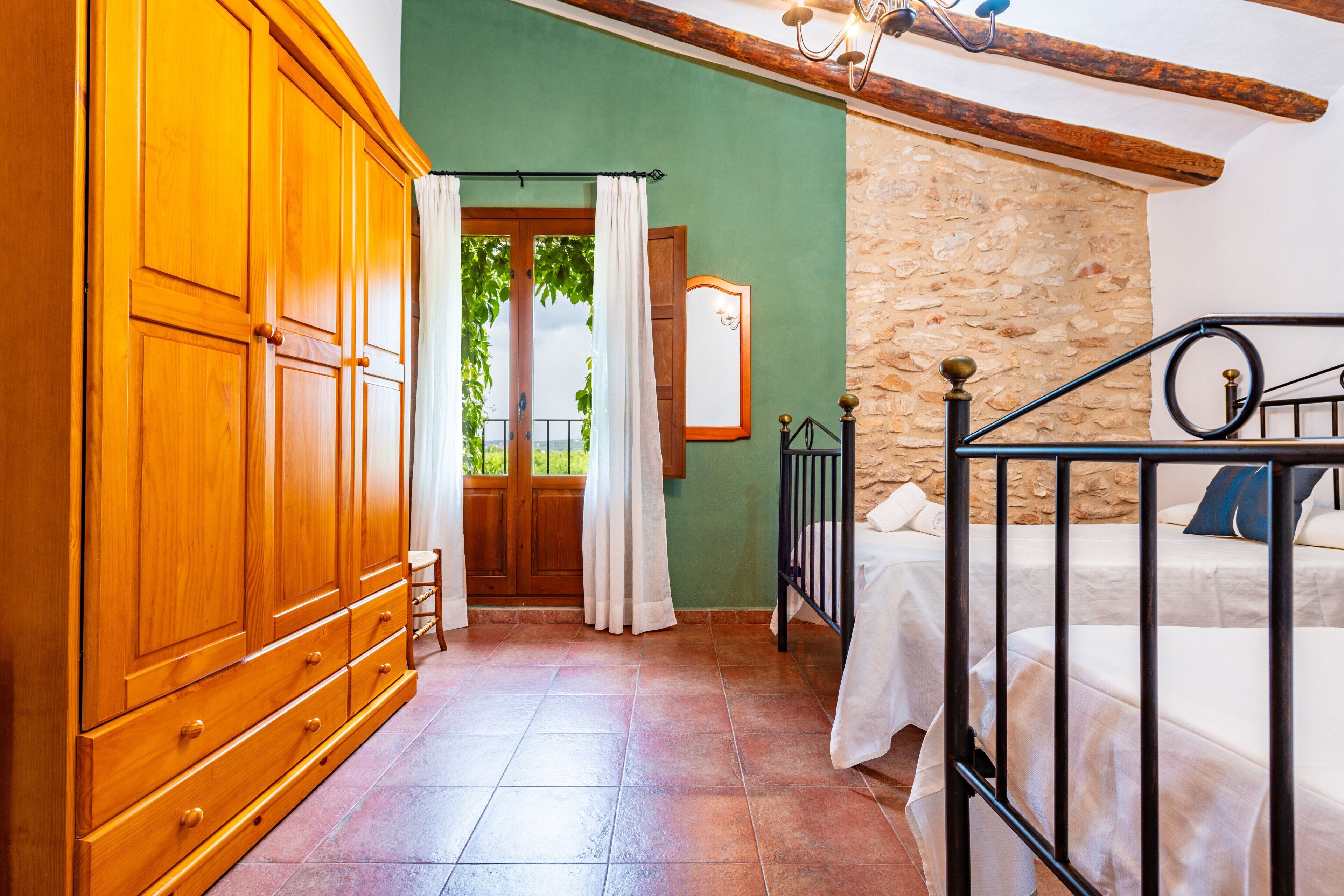 Une chambre avec une armoire en bois, un mur en pierre, un lit avec un cadre en métal et une fenêtre avec des rideaux.