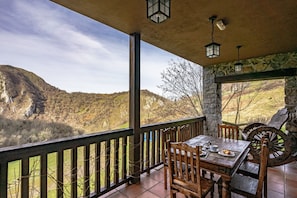 Una mesa de madera con sillas en un balcón, junto a un muro de piedra y vistas a un paisaje montañoso.