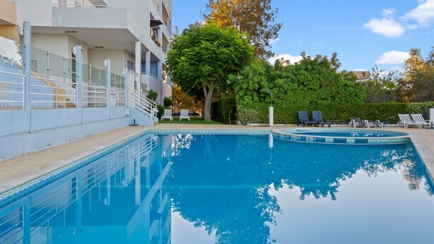 A swimming pool with a clear blue water surface, surrounded by a white building with balconies, a tree, and a hedge. - Apartment 'T1 Em Alvor' with Sea View, Shared Pool and Shared Garden (Alvor)