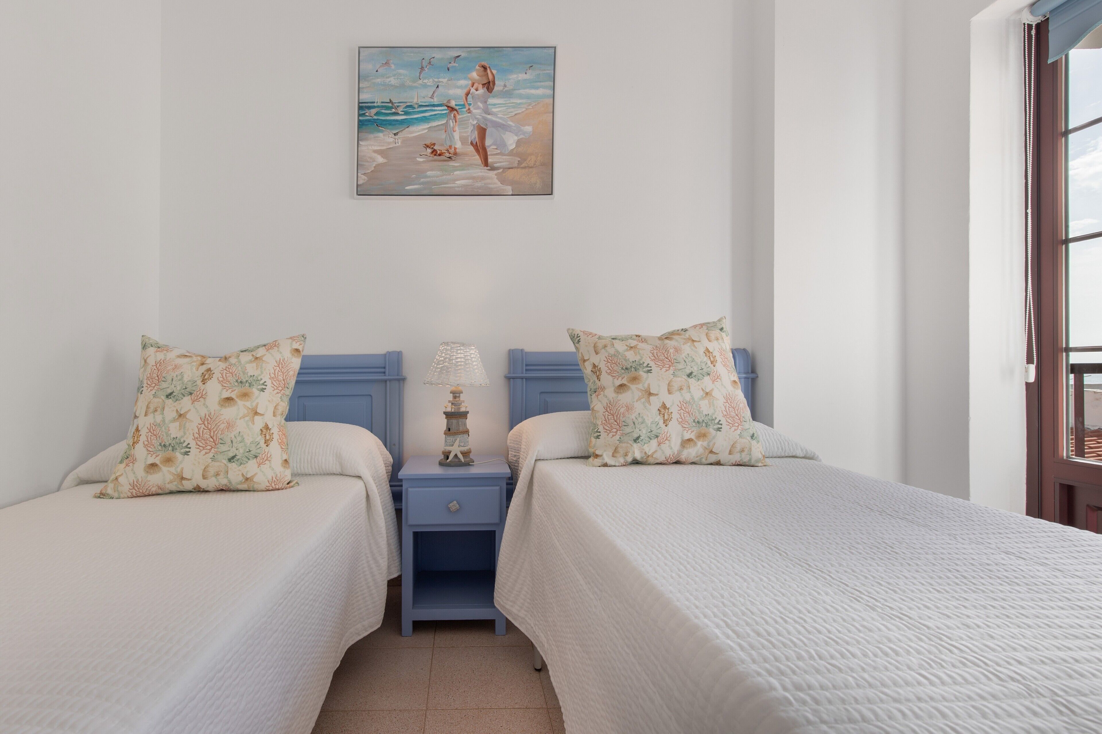 Un lit double agrémenté d’oreillers à motifs floraux, une table de chevet avec une lampe et un tableau représentant une scène de plage.