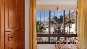A room with a wooden cabinet, a wicker chair with a cushion, and a window with a view of palm trees and buildings.