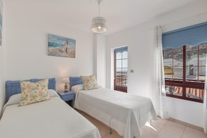 A bedroom with two beds, a painting, a window with a view, and a hanging light fixture.