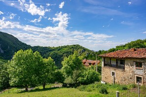 Un bâtiment en pierre surmonté d’un toit de tuiles rouges, entouré d’une végétation luxuriante et adossé à un paysage montagneux.