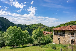 A stone building with a red-tiled roof, surrounded by lush greenery and a mountainous backdrop. - Room 'El Cerezo' with Shared Pool, Shared Garden and Wi-Fi (Gamonedo)
