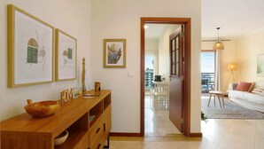 A modern living room with a wooden console table, a large window, and a dining area visible through an open door.