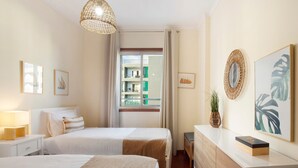 A hotel room with two beds, a bedside table with a lamp, a dresser, a window with a view of buildings, and artwork on the walls.