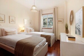 Habitación de hotel con dos camas, una mesita de noche con lámpara, un escritorio con un florero y una ventana con vistas a edificios.