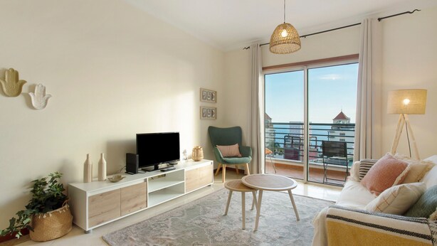 A modern living room with a flat-screen TV, a green armchair, a round coffee table, and a balcony with a view.