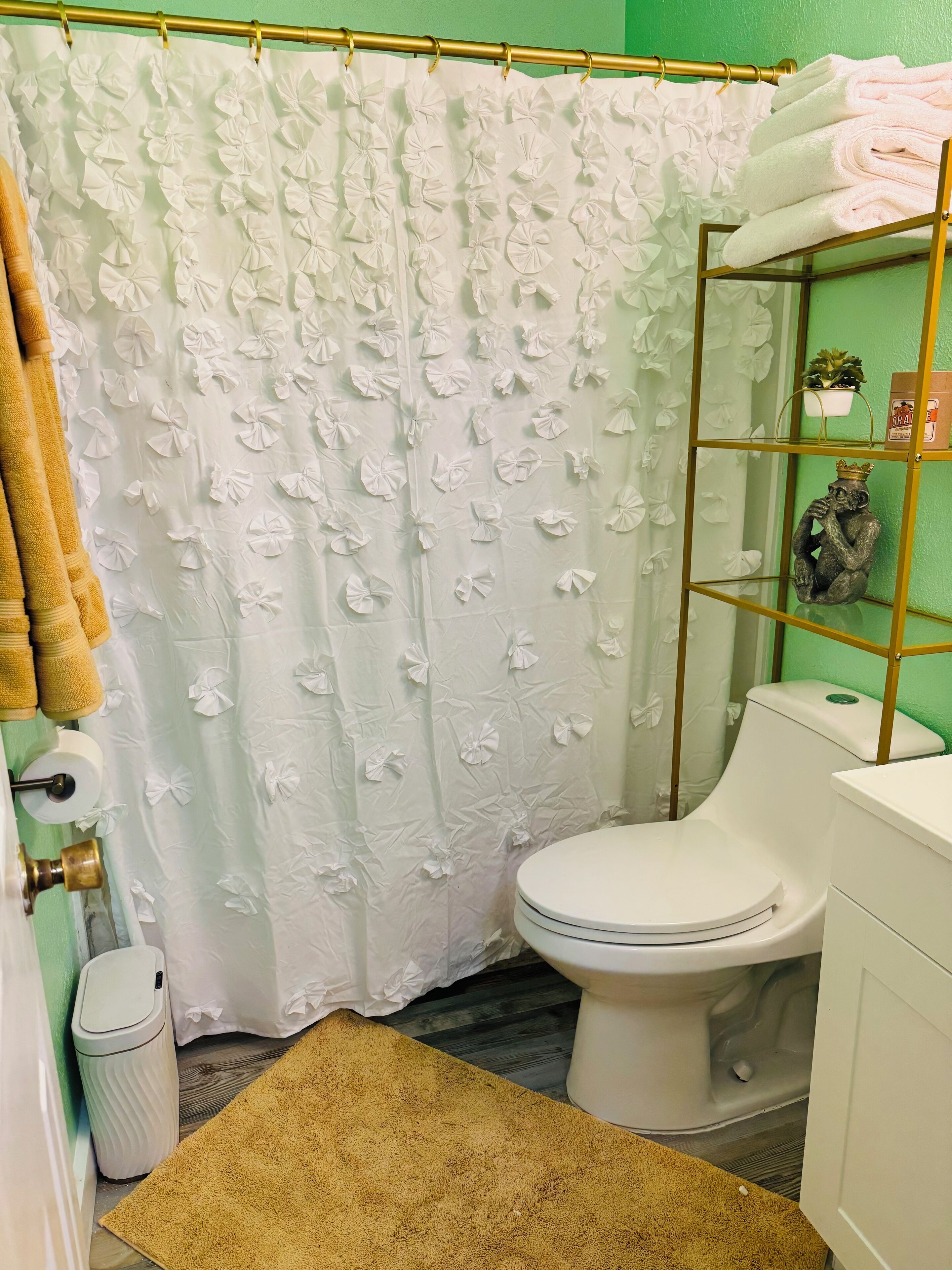 Une salle de bain avec des toilettes blanches, une étagère à cadre doré ornée d’objets décoratifs, un rideau de douche blanc à motifs floraux et un tapis de bain marron.
