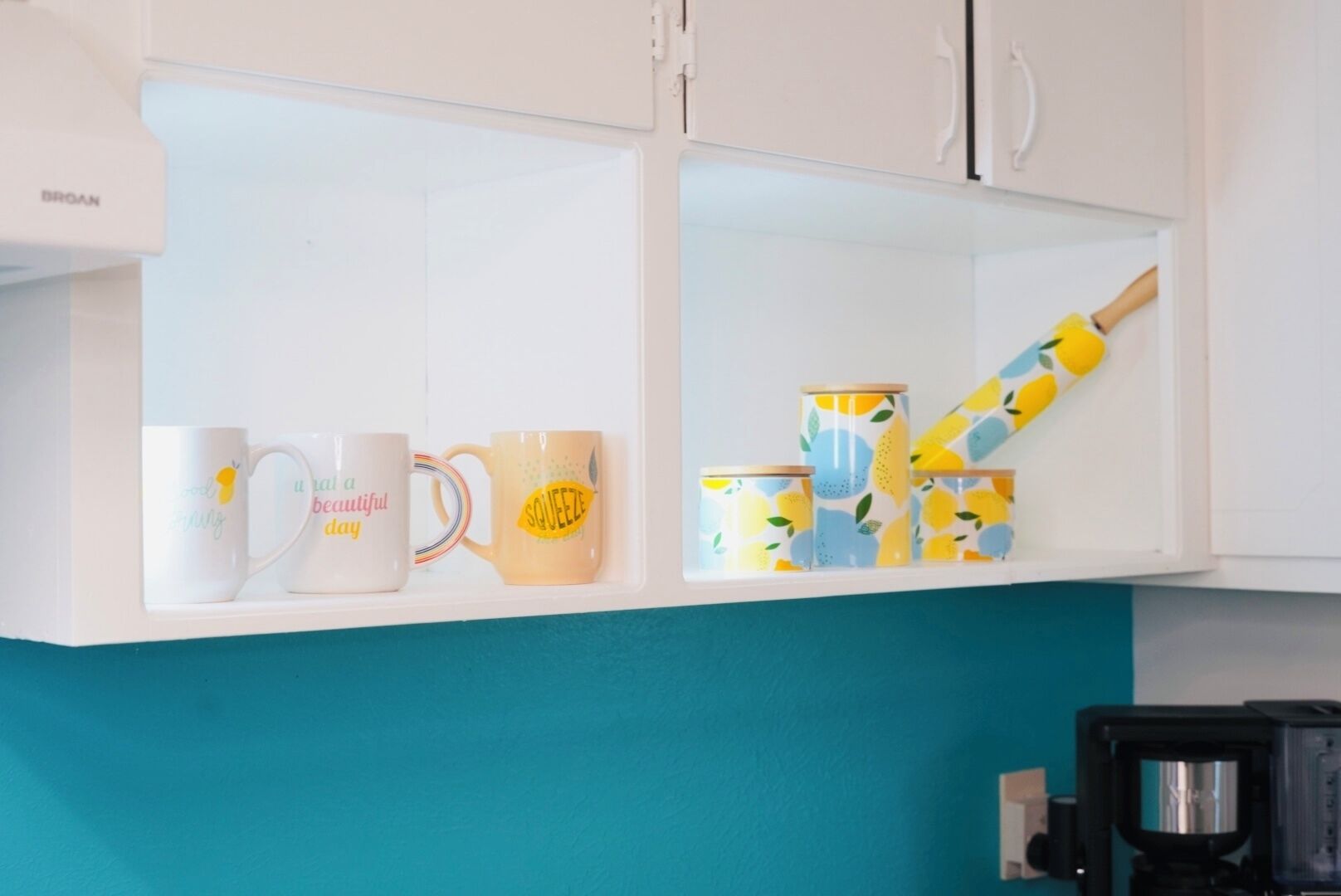 Een keuken met witte kastjes, een blauwgroene spatwand en een koffieapparaat. Op het aanrecht staan drie mokken, elk met verschillende designs en tekst.
