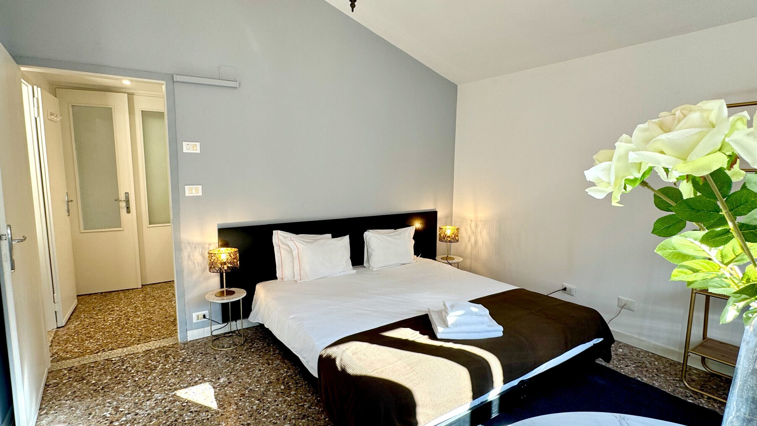 Camera d'hotel con un letto grande, due abat-jour, un comodino, un vaso con fiori bianchi e una porta a sinistra.