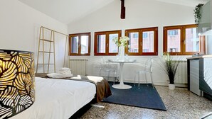 A hotel room with a bed, a desk, a chair, a radiator, and windows.