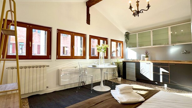 A hotel room with a dining table, chairs, a radiator, a chandelier, and a view of the outside through large windows.