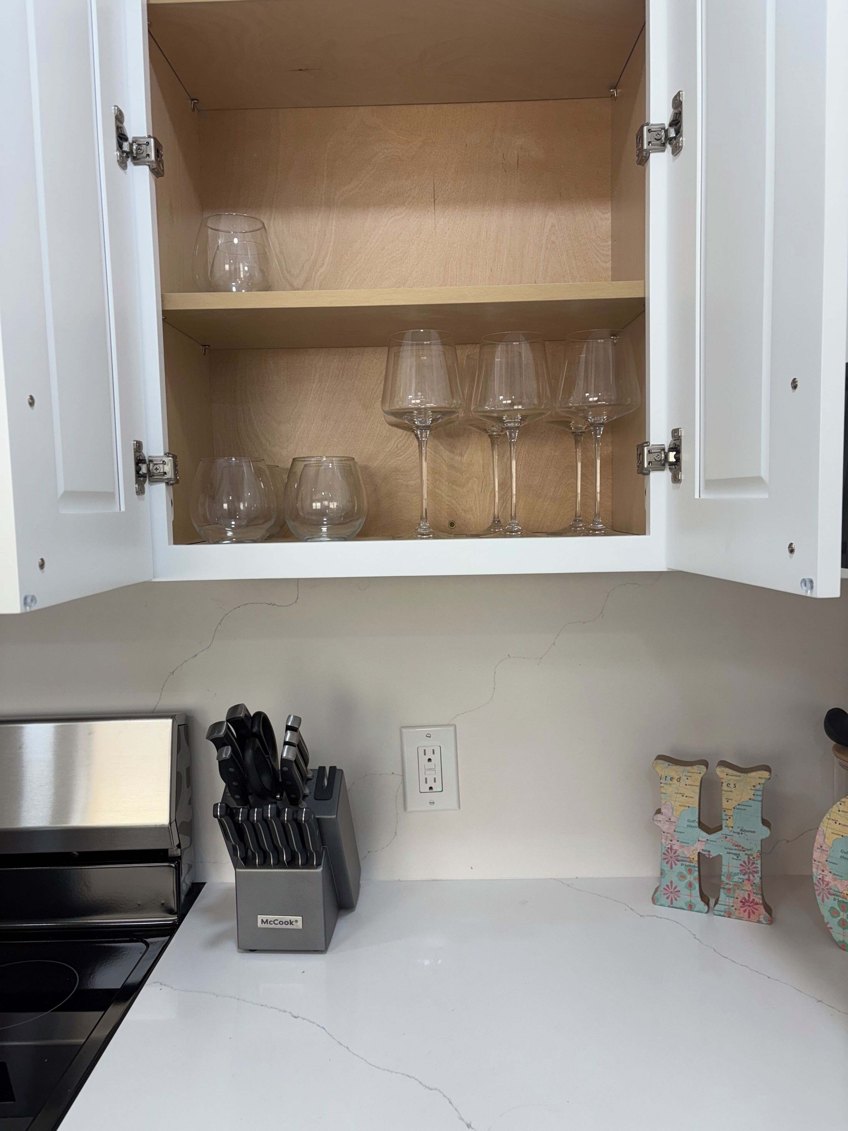 Une cuisine avec une étagère en bois où sont rangés des verres à vin, un bloc à couteaux McCook et une lettre décorative « H » accrochée au mur.