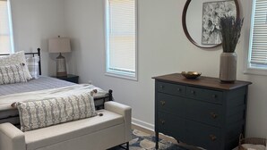 A bedroom with a bed, dresser, bedside table, lamp, and a large window with blinds.