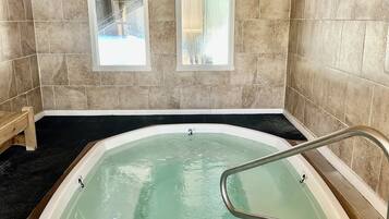 A hot tub with a wooden ceiling and stone walls, equipped with two windows and a metal ladder.