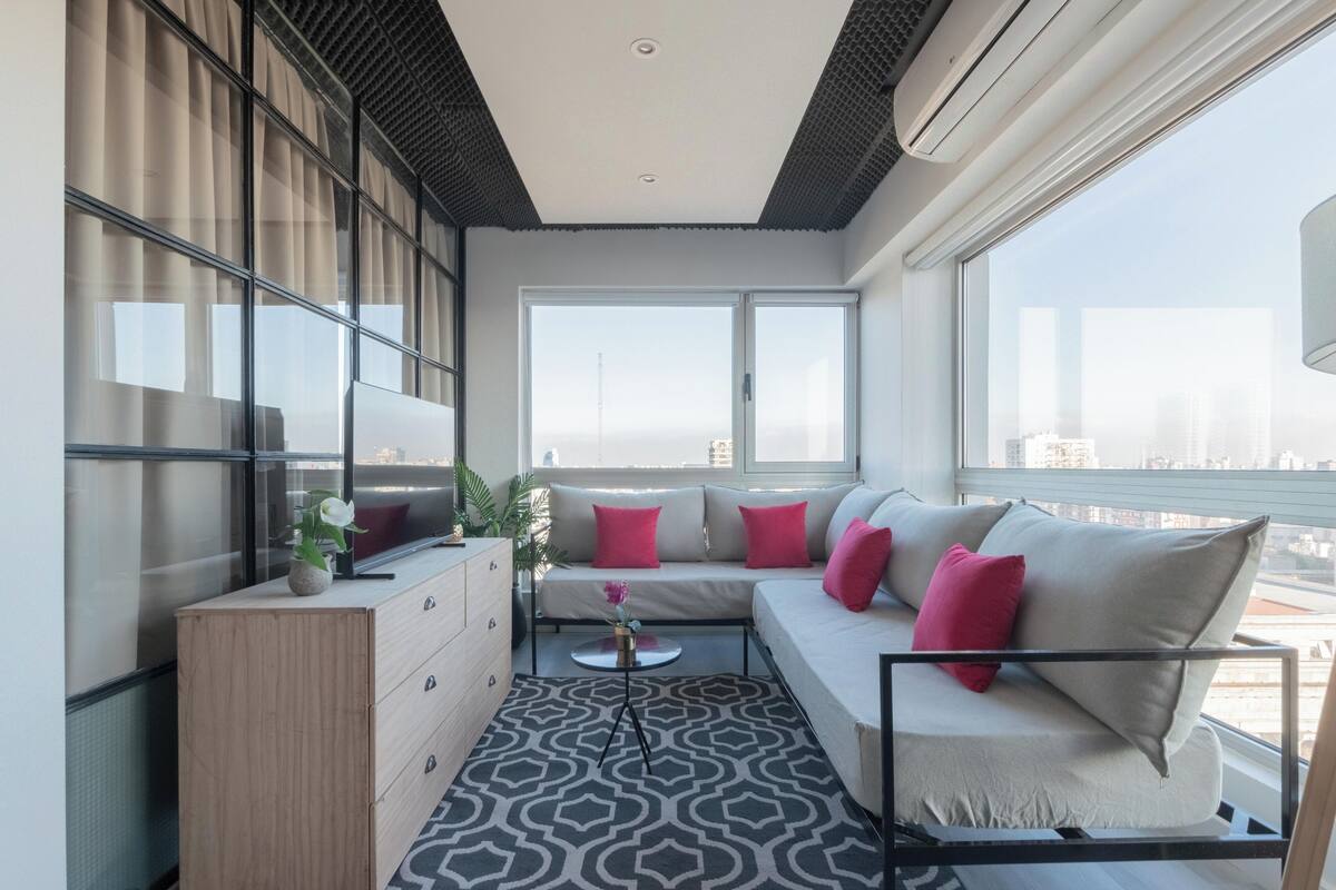 A modern living room with a sofa, a coffee table, and a dresser. There are large windows offering a view of the city.