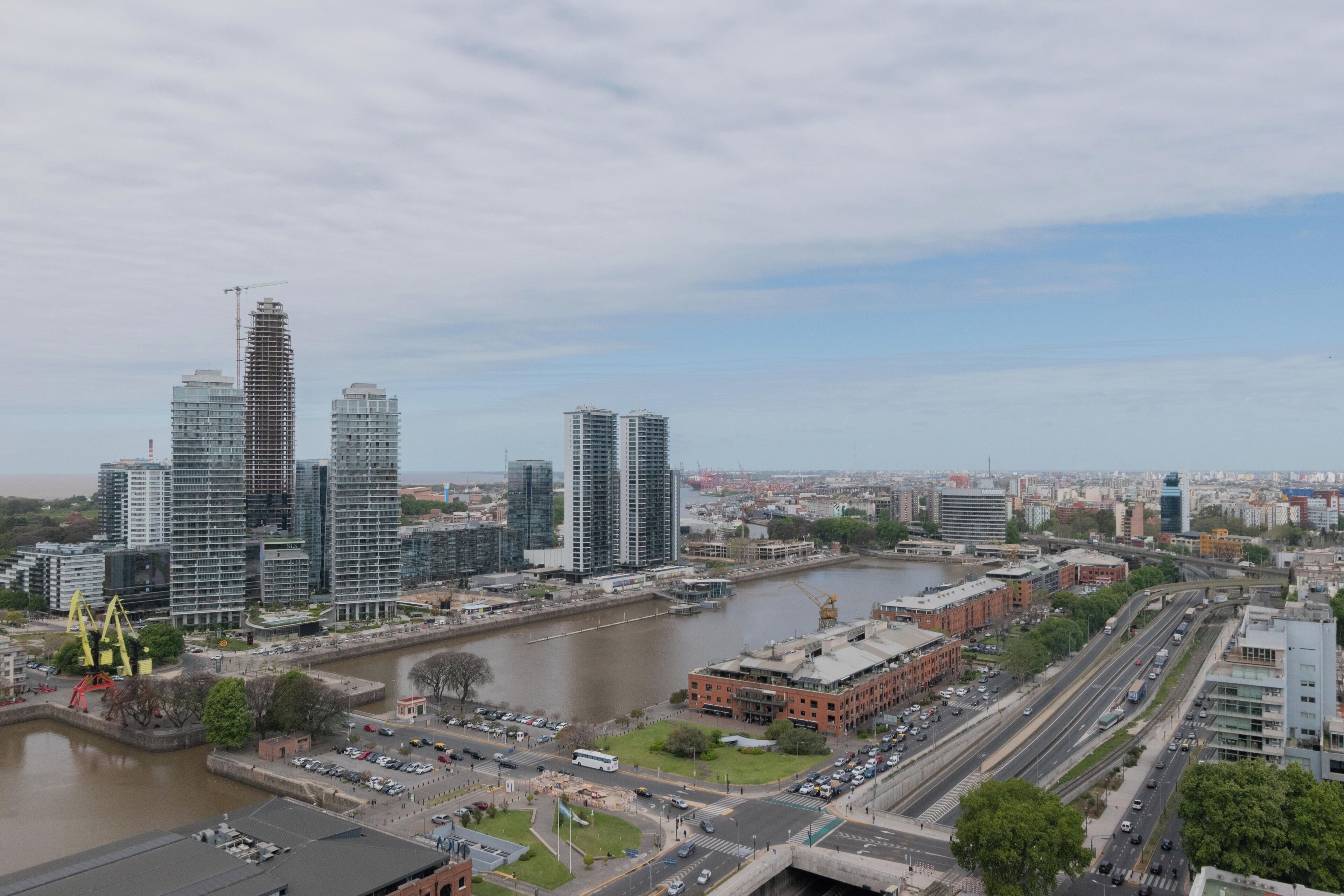 Un paysage urbain avec des immeubles modernes de grande hauteur, une rivière et une rue animée où circulent de nombreux véhicules.