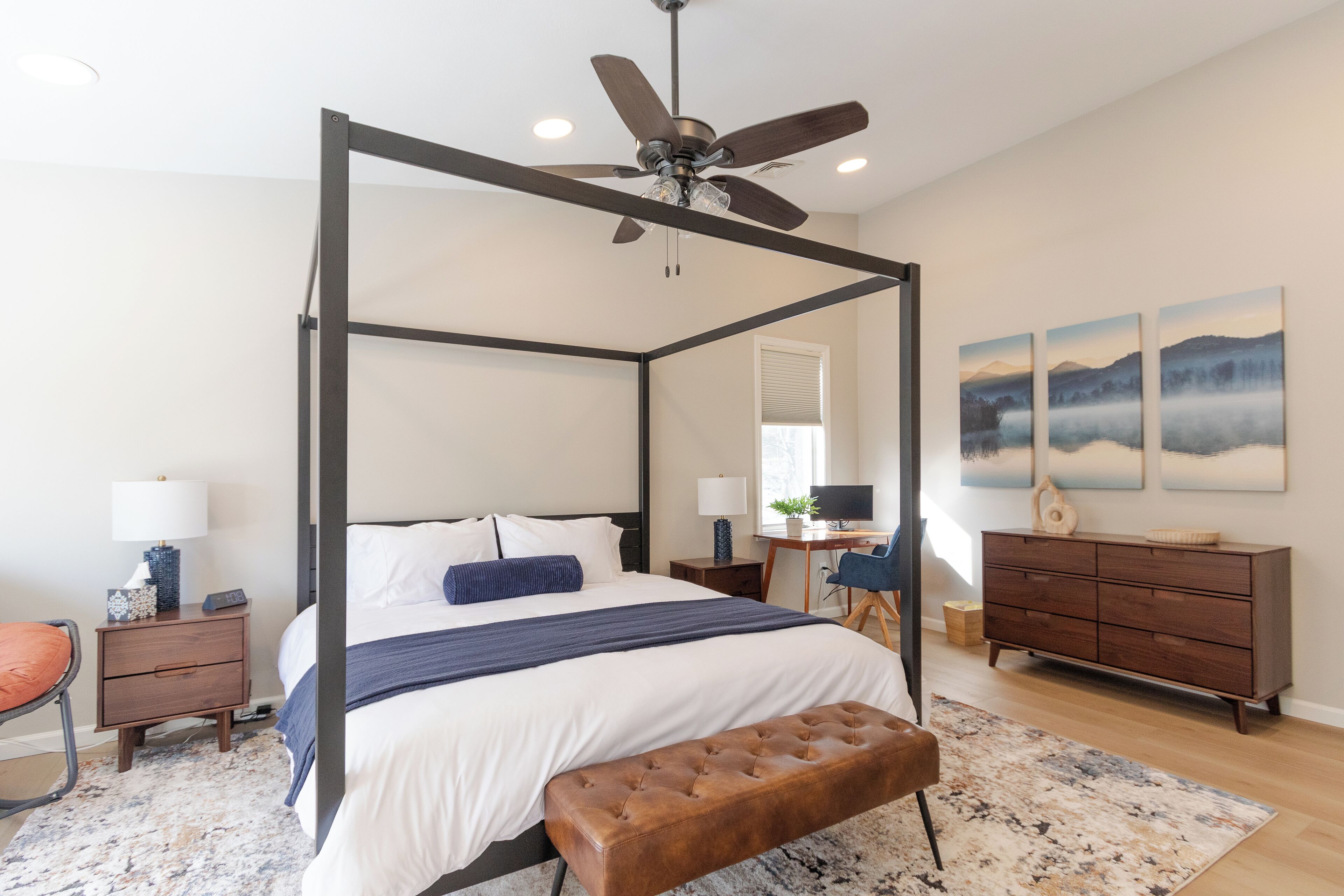 Un lit à baldaquin à quatre poteaux avec un ventilateur de plafond, un bureau avec un ordinateur, une commode et des œuvres d’art accrochées au mur.