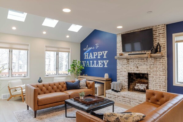 Een woonkamer met een open haard, een grote tv erboven bevestigd, en een muur met een bord waarop 'Welcome to Happy Valley' staat.