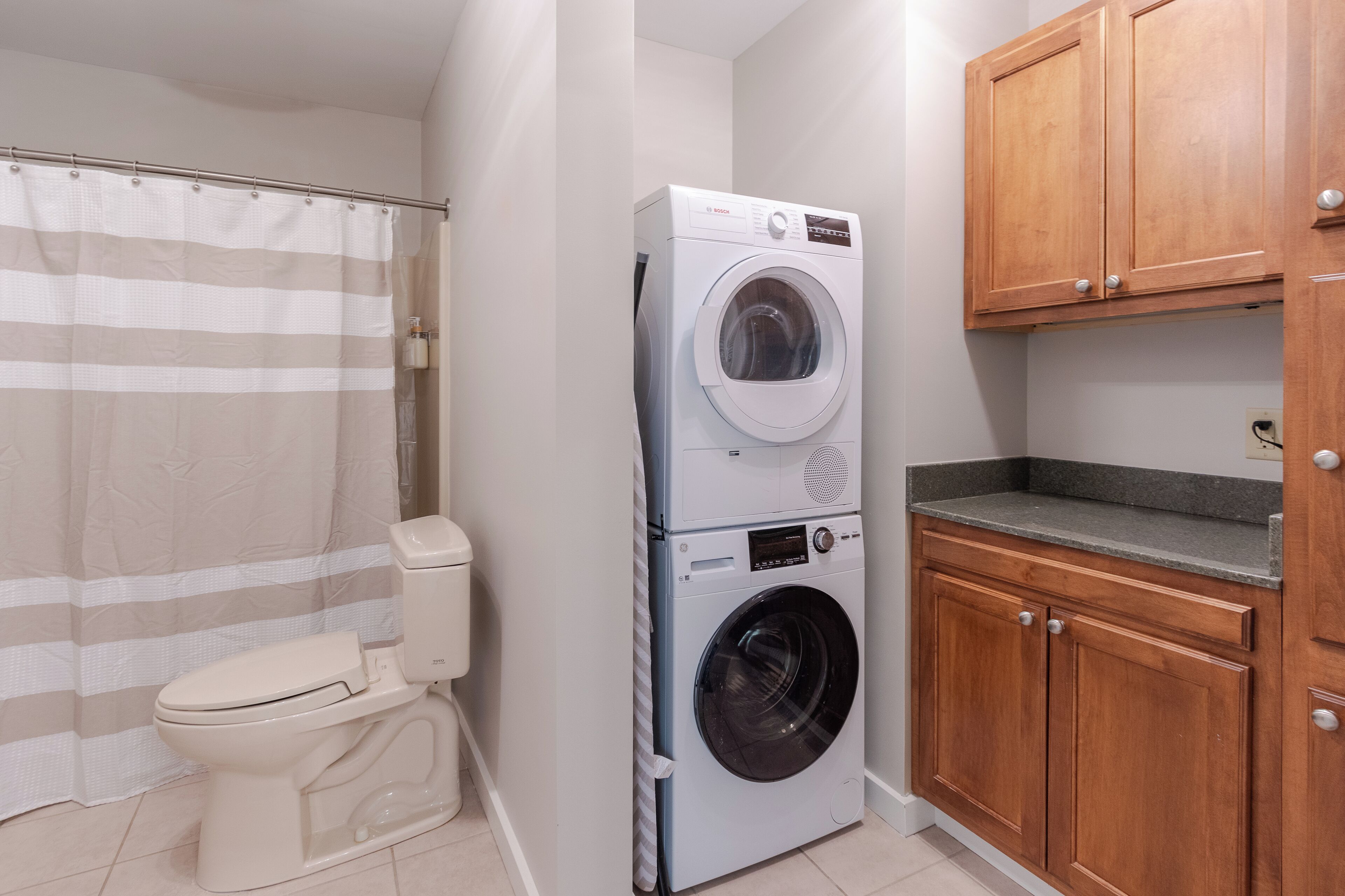 A compact bathroom with a white toilet, a stacked washer and dryer, a shower curtain, and wooden cabinets.