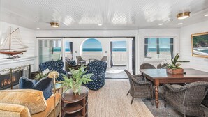 A spacious living room with a view of the beach, featuring a fireplace, comfortable seating, and a dining area. - Luxury Oceanfront Condo - Perfect for Large Families - Steps to the Sand! (Newport Beach)