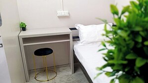 A hotel room with a bed, a desk with a chair, a small plant, and a power outlet on the wall.