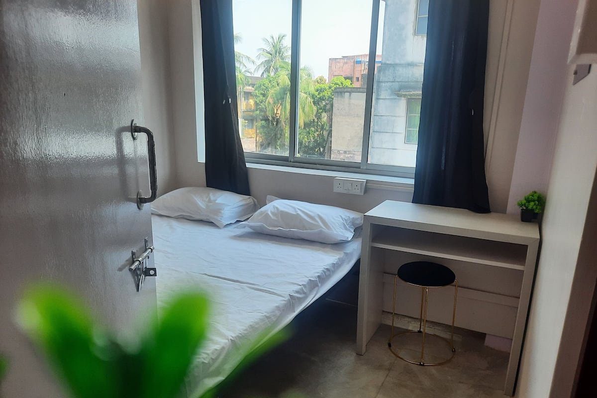 A small, clean room with a bed, a desk, and a window overlooking a building.