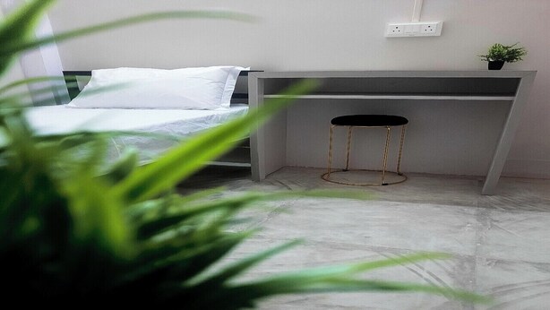 A bedroom with a bed, a desk, and a chair. There is a plant in the foreground.