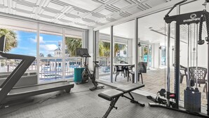 Un gimnasio con equipo de ejercicio, ventanas amplias y vistas a una piscina y palmeras.