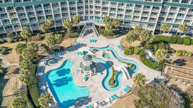 Vista aérea de un complejo turístico con un amplio área de piscina, varias toboganes de agua y un edificio de varios pisos al fondo.