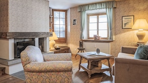 Ein gemütliches Wohnzimmer mit Kamin, floralen Ohrensesseln, einem Holztisch und Blick ins Freie durch ein Fenster.