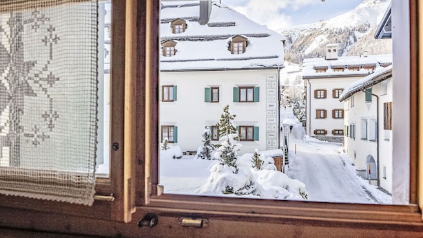 Blick aus dem Fenster auf eine verschneite Straße mit Gebäuden, einer Straßenlaterne und einem Berg im Hintergrund.