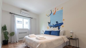 A bedroom with a large bed, white bedding, a wall mural of fish, a potted plant, and a small table with a lamp. - MOT Charcas 3000 (Buenos Aires)