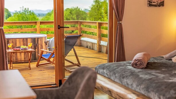 Une chambre avec un lit, une chaise, une table sur laquelle se trouve un saladier de fruits, et une vue sur une terrasse en bois et la verdure environnante.