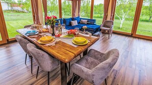 Un intérieur en bois avec une table à manger dressée pour quatre personnes, un canapé bleu et une vue sur un jardin verdoyant, visible à travers de grandes fenêtres.