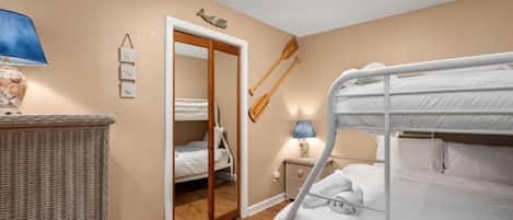 Una habitación con literas, una mesita de mimbre, una mesita de noche con lámpara, y una puerta que da a otra habitación.