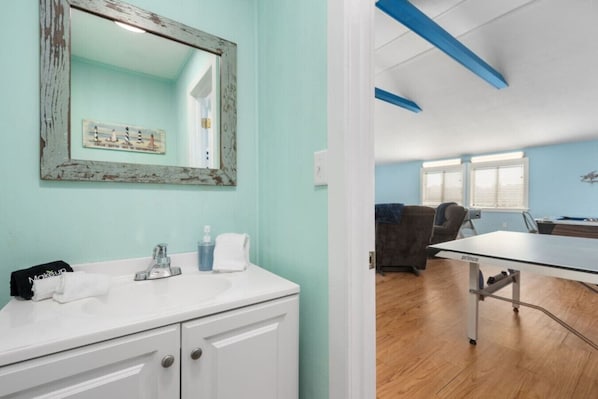 A room with a white vanity, a mirror, a framed picture, a table tennis table, and a seating area with a couch and chairs.
