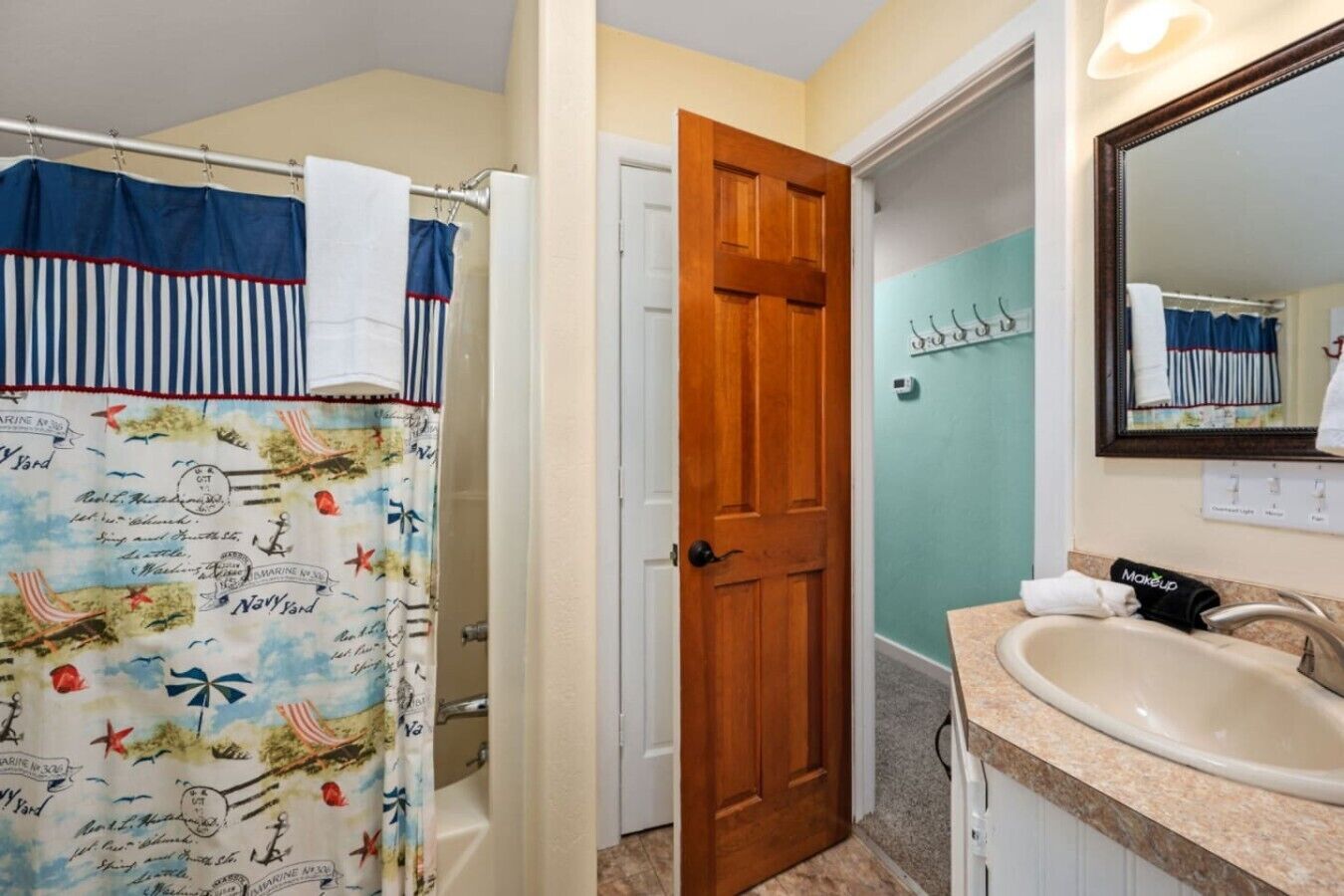 A bathroom with a shower curtain featuring maritime designs, a wooden door, a sink with a makeup towel, and a mirror.