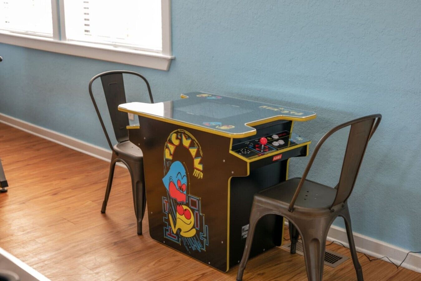 Um quarto com piso de madeira, duas cadeiras, uma mesa pequena com um aparelho de jogos e uma janela com persianas.