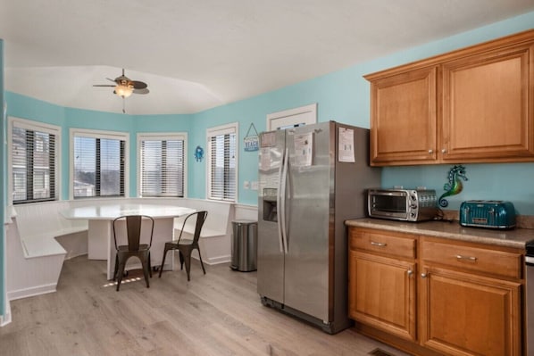 A kitchen with a refrigerator, microwave, and toaster, wooden cabinets, a white countertop, and a dining area with a table and chairs.
